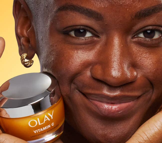Person smiling, holding Olay Vitamin G Lightweight Moisturizer adjacent to their look with a lukewarm backdrop