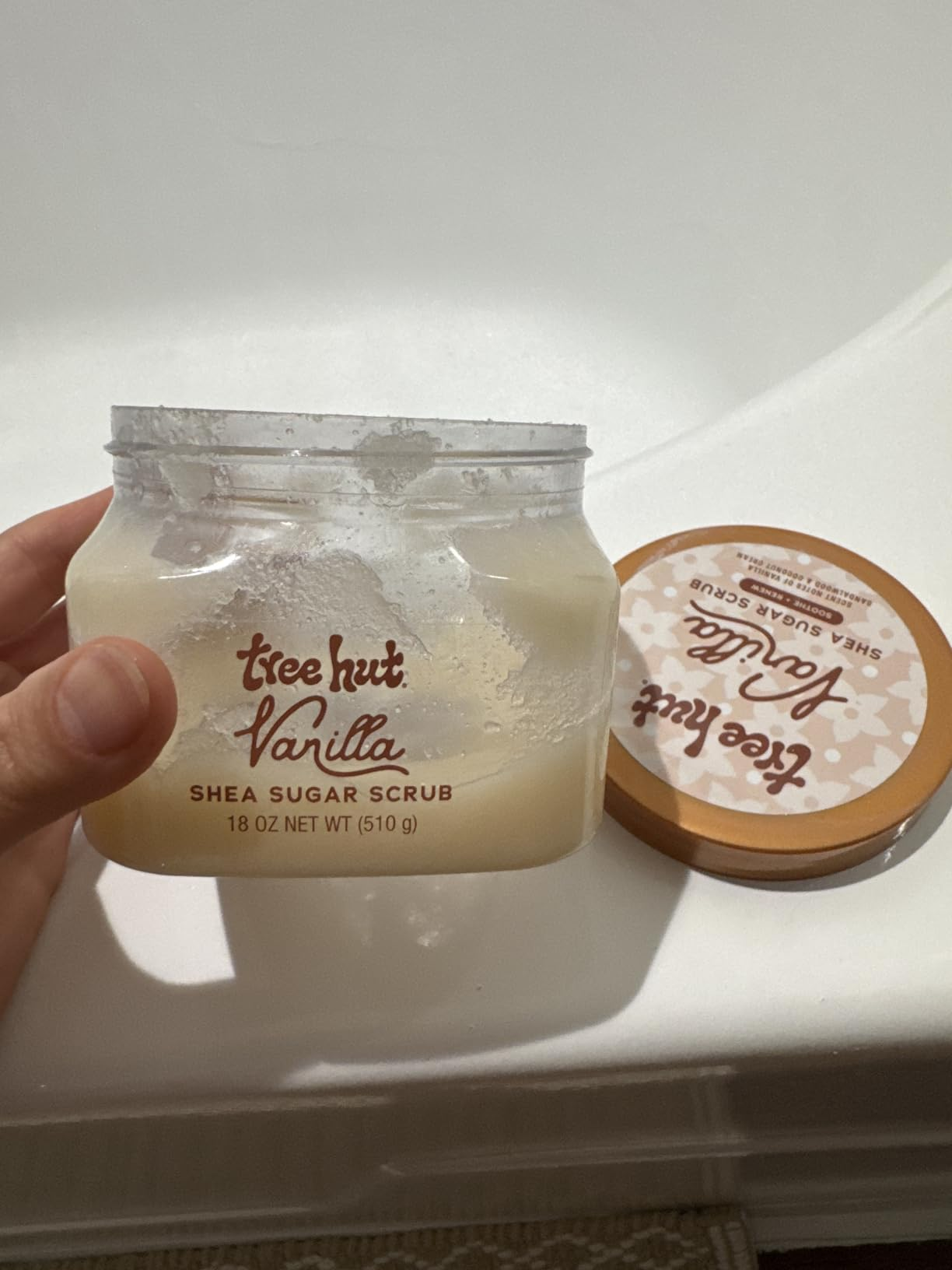 A manus  holds an unfastened  jar of Tree Hut Vanilla Shea Sugar Scrub, showing its creamy texture. The lid is placed beside the jar
