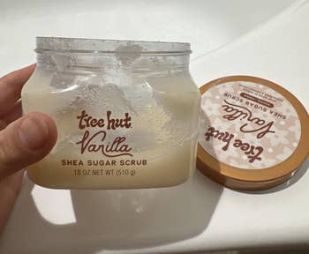 A manus holds an unfastened jar of Tree Hut Vanilla Shea Sugar Scrub, showing its creamy texture. The lid is placed beside the jar