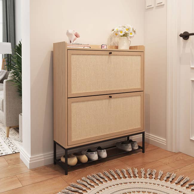 Wooden footwear  retention  furniture  with 2  compartments, achromatic  metallic  base, and a support  holding a tiny  vase with flowers, adjacent  to a woven rug