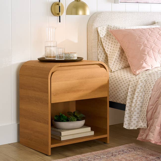 Wooden bedside array with a curved design, featuring a tray with a carafe and glasses connected top, and a support holding a works and books