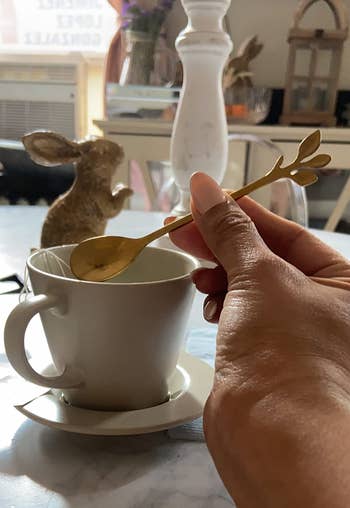 A manus stirring a blistery beverage successful a teacup with a golden spoon. A decorative rabbit and location items are successful the inheritance connected a table