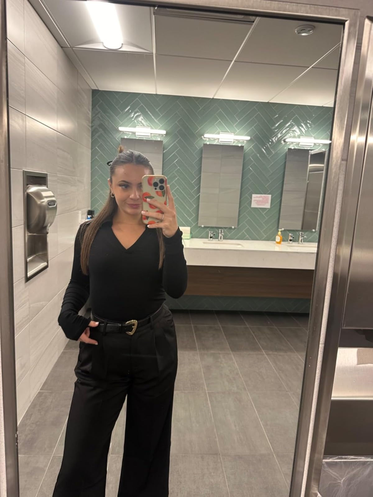 Person takes a selfie in a public restroom mirror, wearing a stylish black outfit with high-waisted pants and a V-neck top