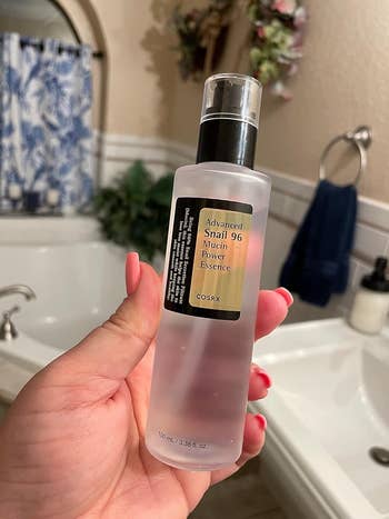 Tangan memegang sebotol COSRX COSRX Lanjutan Siput 96 Mucin Power Essence dalam pengaturan kamar mandi