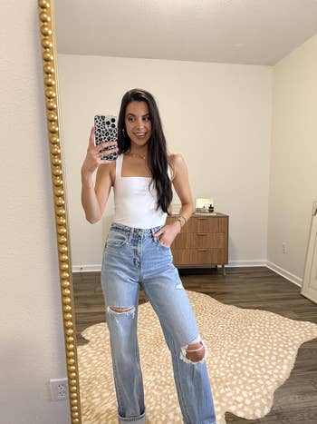 Woman taking a mirror selfie wearing a white sleeveless top and ripped jeans, standing on a patterned rug in a modern room