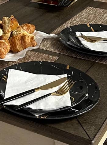 Elegant dining table set with black plates, black and gold flatware, and croissants