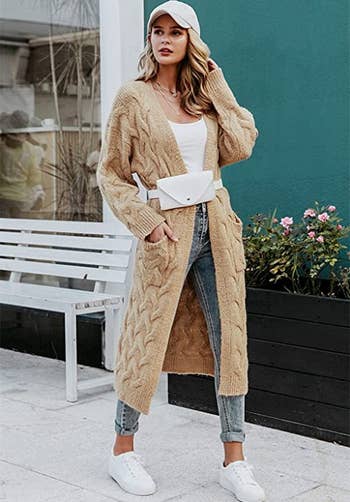a model wearing a long tan knit cardigan over a white shirt and paired with blue jeans and white sneakers