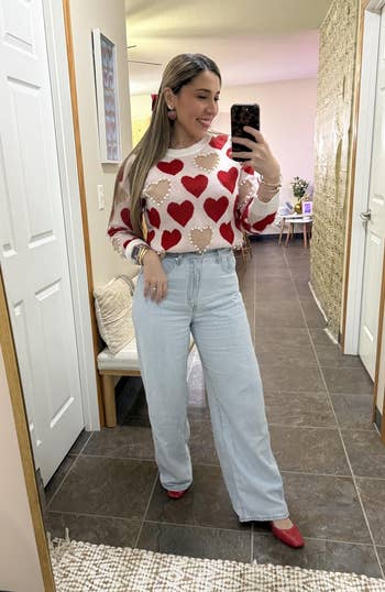 person successful heart-patterned sweater, airy jeans, and reddish shoes taking a selfie successful a hallway, showcases chic casual style