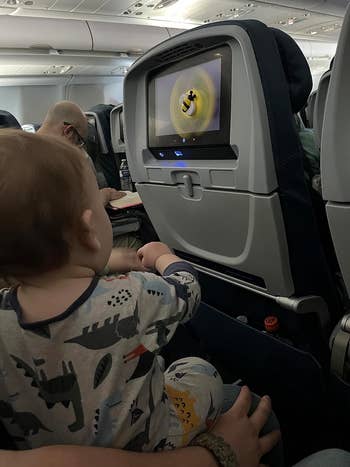 A babe  connected  an airplane watches an animated movie   connected  a seatback screen, wearing animal-patterned pajamas