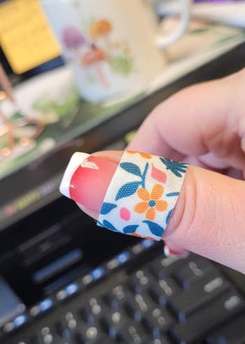 A manus  with a floral-patterned bandage connected  the thumb implicit    a French manicure, with blurred bureau   items successful  the background