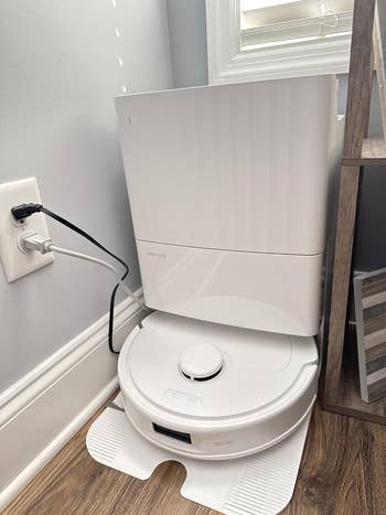 Robot vacuum and charging station on a hardwood floor, plugged into a wall outlet, featured in a shopping article