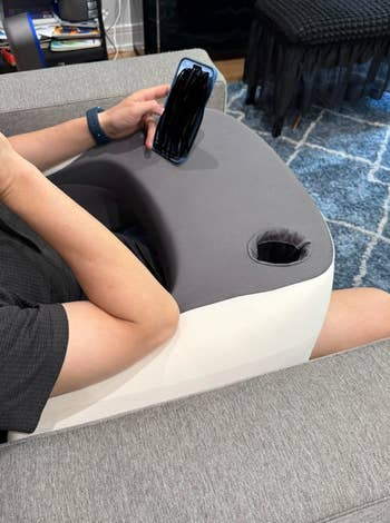 Person utilizing a gray, wedge-shaped pillow tray with a built-in cupful  holder, sitting connected  a couch, holding a smartphone
