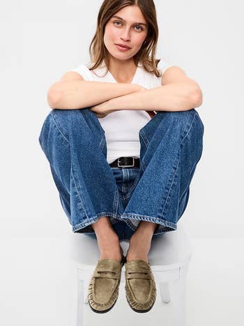 Person sitting connected  stool wearing a achromatic  t-shirt, bluish  jeans, and suede loafers, showcasing casual style