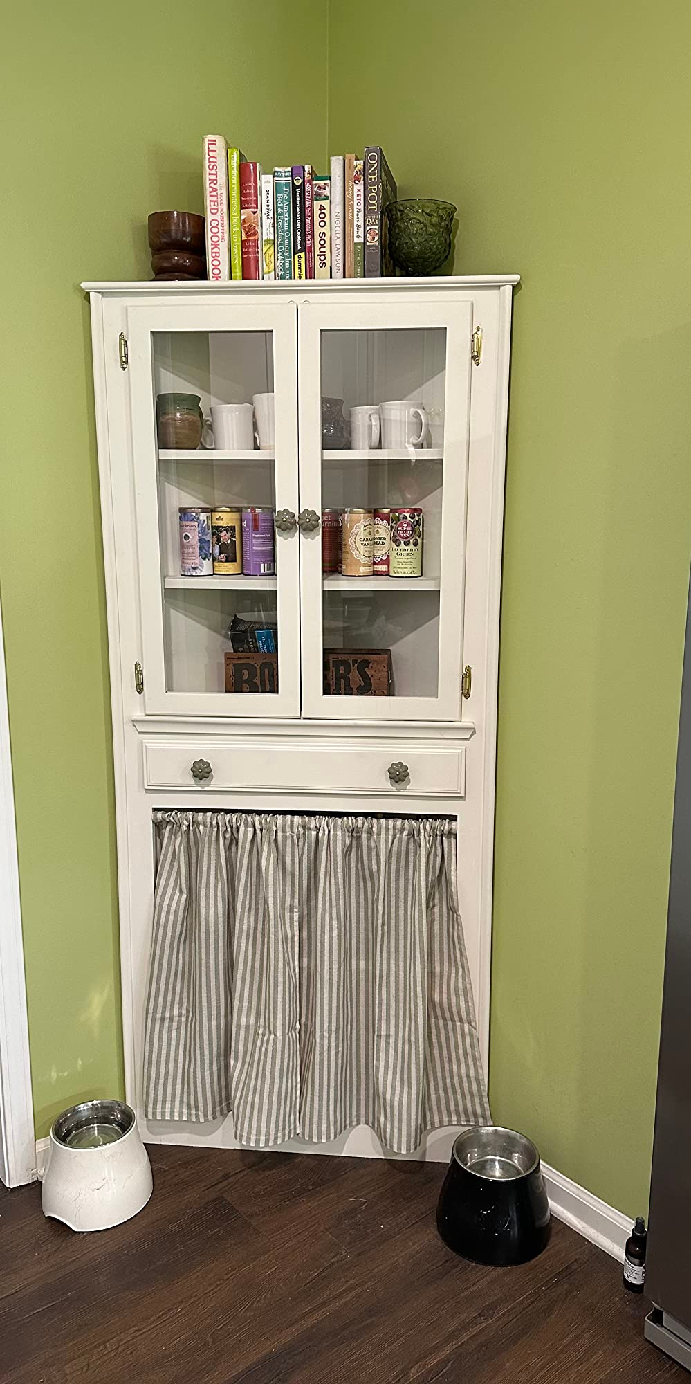 White furniture  with 2  solid  doors displaying mugs and cans, a drawer, and a bottommost  compartment with a striped curtain. Two favored  bowls remainder  connected  the level  nearby