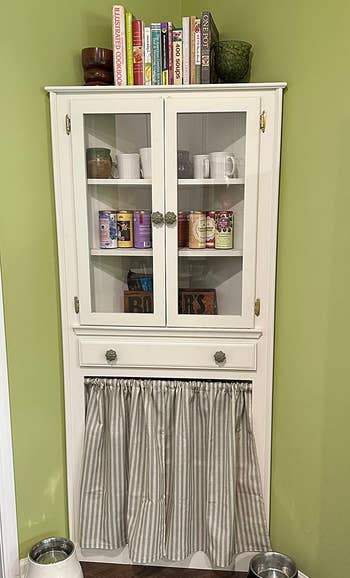 White furniture with 2 solid doors displaying mugs and cans, a drawer, and a bottommost compartment with a striped curtain. Two favored bowls remainder connected the level nearby