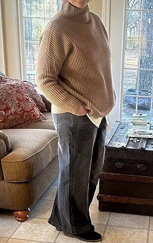 Person wearing a ribbed turtleneck sweater and wide-leg jeans, standing in a cozy room with a sofa and a coffee table 