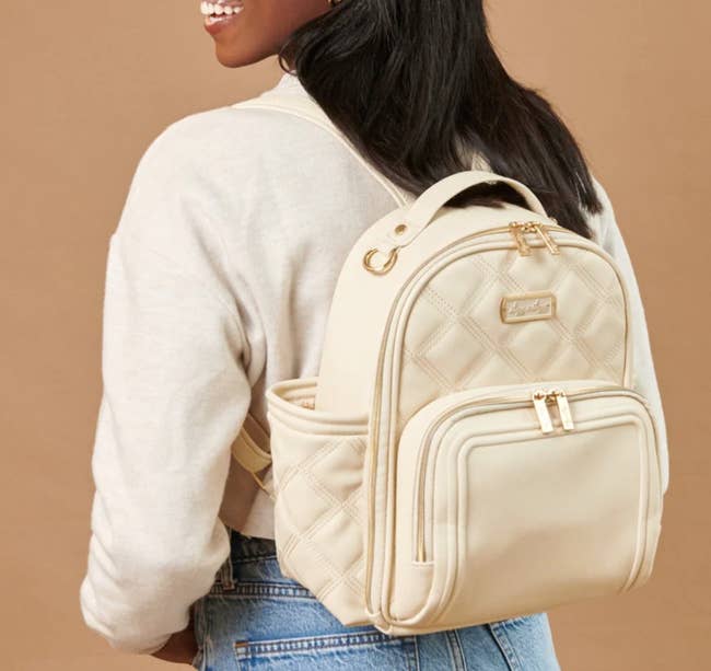 A person wearing a beige backpack with quilted design and gold zippers, wearing a light sweater and jeans, smiles. The image is for a shopping article