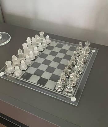 A clear glass chess set arranged for the start of a game on a dark table