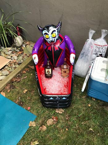 Inflatable vampire cooler holding 2  bottles, filled with ice, sits connected  grass. Nearby are bags of crystal  and a cooler, suggesting an outdoor gathering