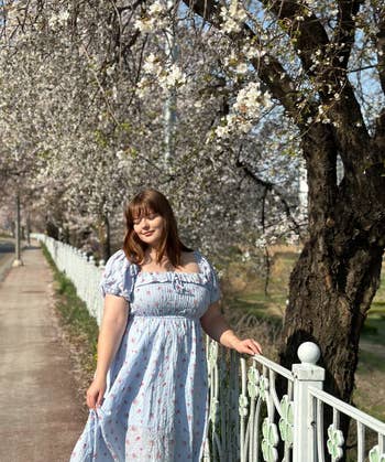 Woman in a breezy floral dress stands by a fence with blossoming trees in the background. Ideal spring fashion
