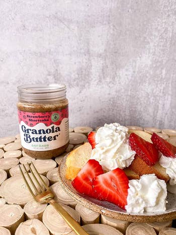 Jar of Strawberry Shortcake Granola Butter next to toast with cream and sliced strawberries on a plate