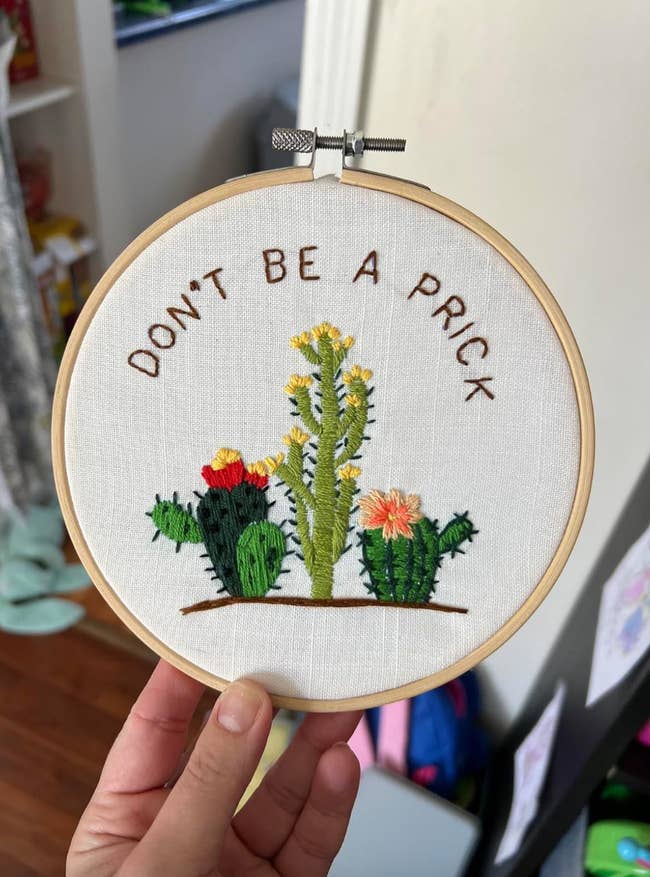 A hand holds a circular embroidery hoop displaying a design with three cacti and the text 