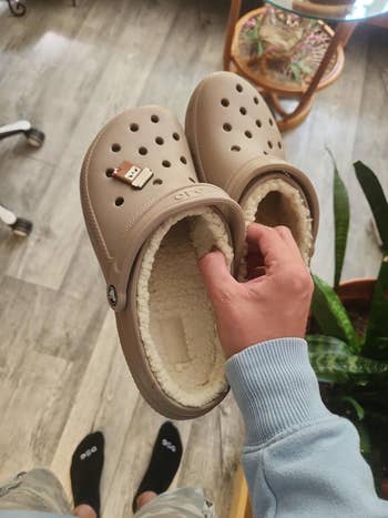 Person holding a pair of beige fuzzy-lined Crocs with a coffee cup charm, standing indoors on a wood floor next to a chair and plants