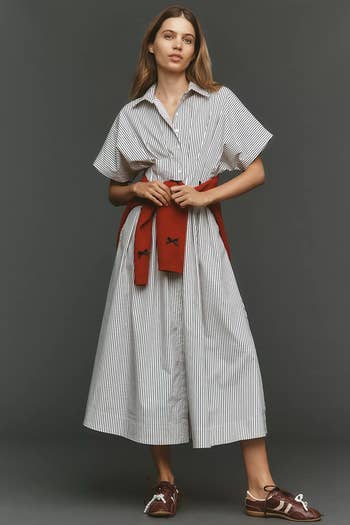 Model wearing a striped shirt dress with a red sweater tied at the waist and casual brown shoes. Fashionable, relaxed style for shopping