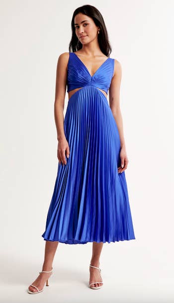 Woman models a pleated blue midi dress with a V-neckline and thin straps, paired with simple heels
