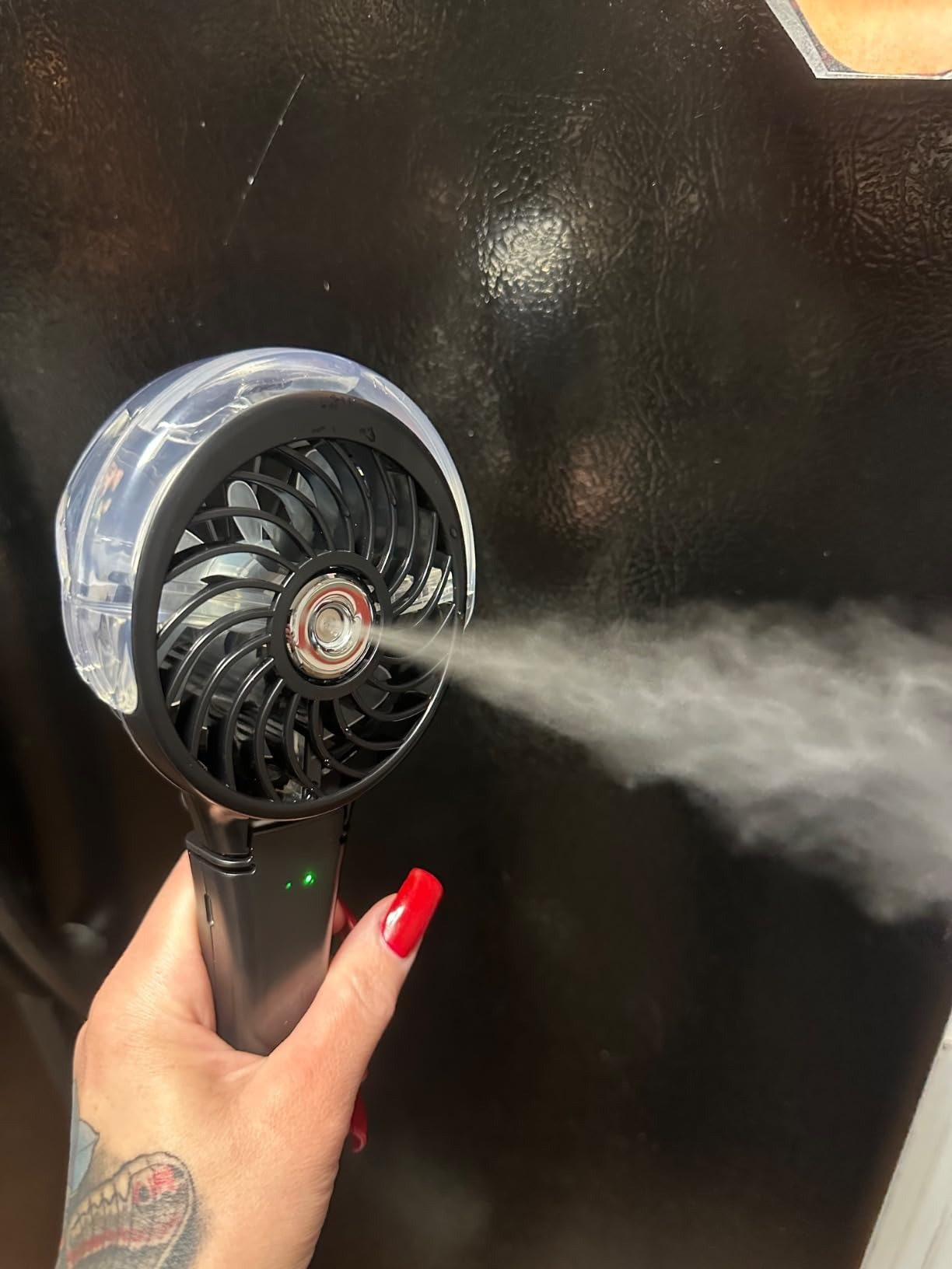 Hand holding a portable misting fan, emitting good  mist; idiosyncratic    has reddish  nail polish and a tattoo