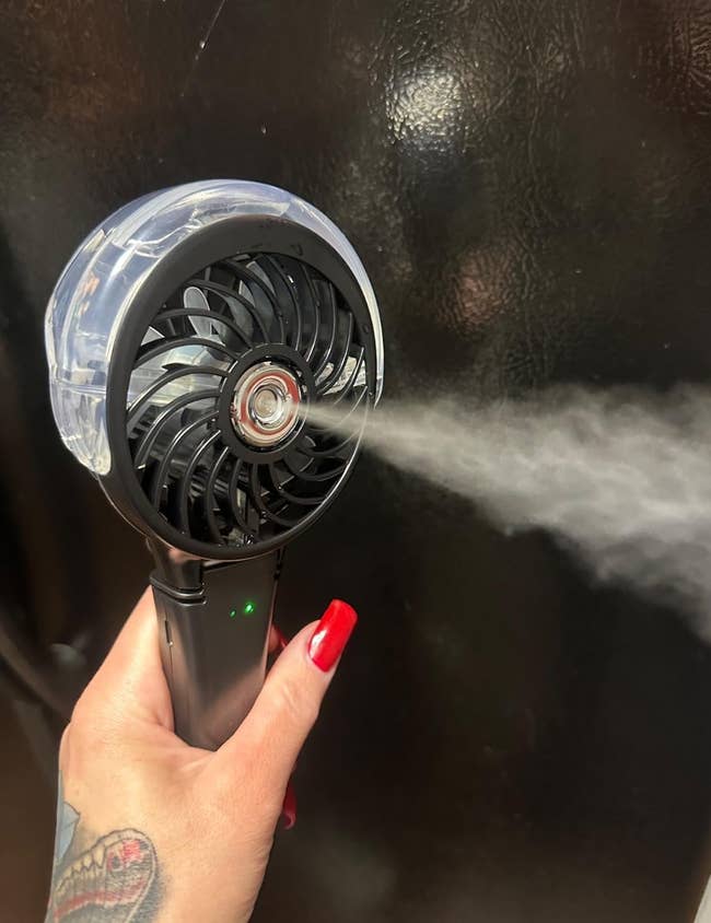 Hand holding a portable misting fan, emitting good mist; idiosyncratic has reddish nail polish and a tattoo
