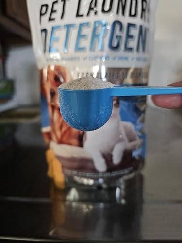 A scoop of pet laundry detergent powder is held in front of its packaging, which features a dog and a cat image