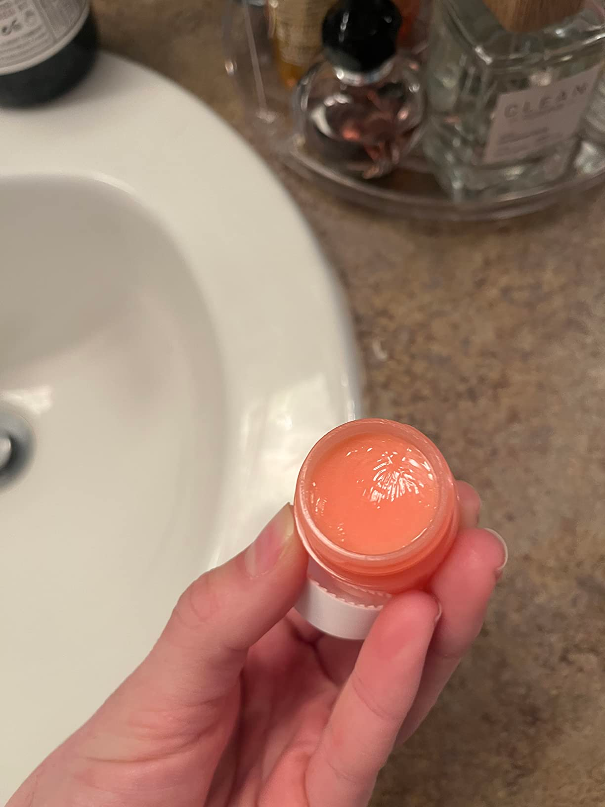 A hand holding an open jar of lip balm near a bathroom sink, with skincare products in the background