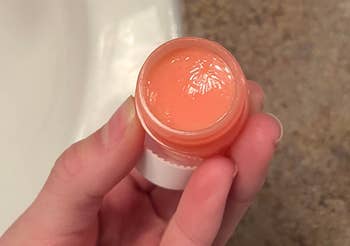 A hand holding an open jar of lip balm near a bathroom sink, with skincare products in the background