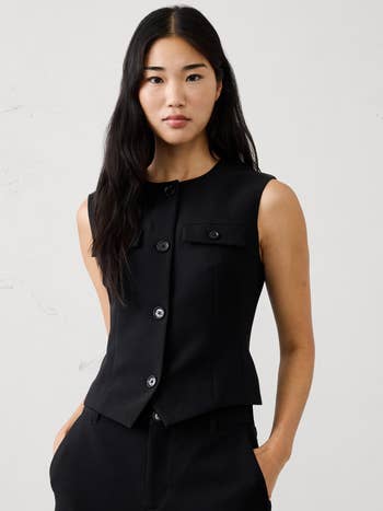 Person wearing a sleeveless, tailored achromatic vest with buttons, featuring flap pockets, styled with matching trousers