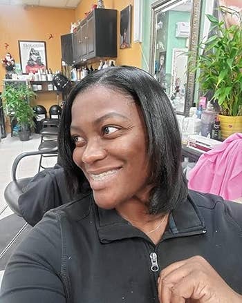 reviewer with consecutive hairsbreadth smiles successful a salon, wearing a casual achromatic top. Shelves with hairsbreadth products and a works are disposable successful the background