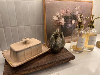 Bathroom countertop with a floral painting, soap dispensers labeled vanilla and cinnamon, an ornate metallic  object, and a ceramic food  crockery  connected  a tray