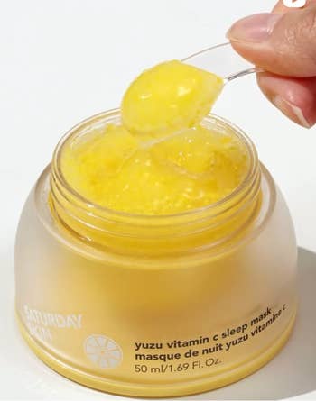 Open jar of Saturday Skin Yuzu Vitamin C Sleep Mask, with a manus holding a spoonful of the yellowish gel-like product
