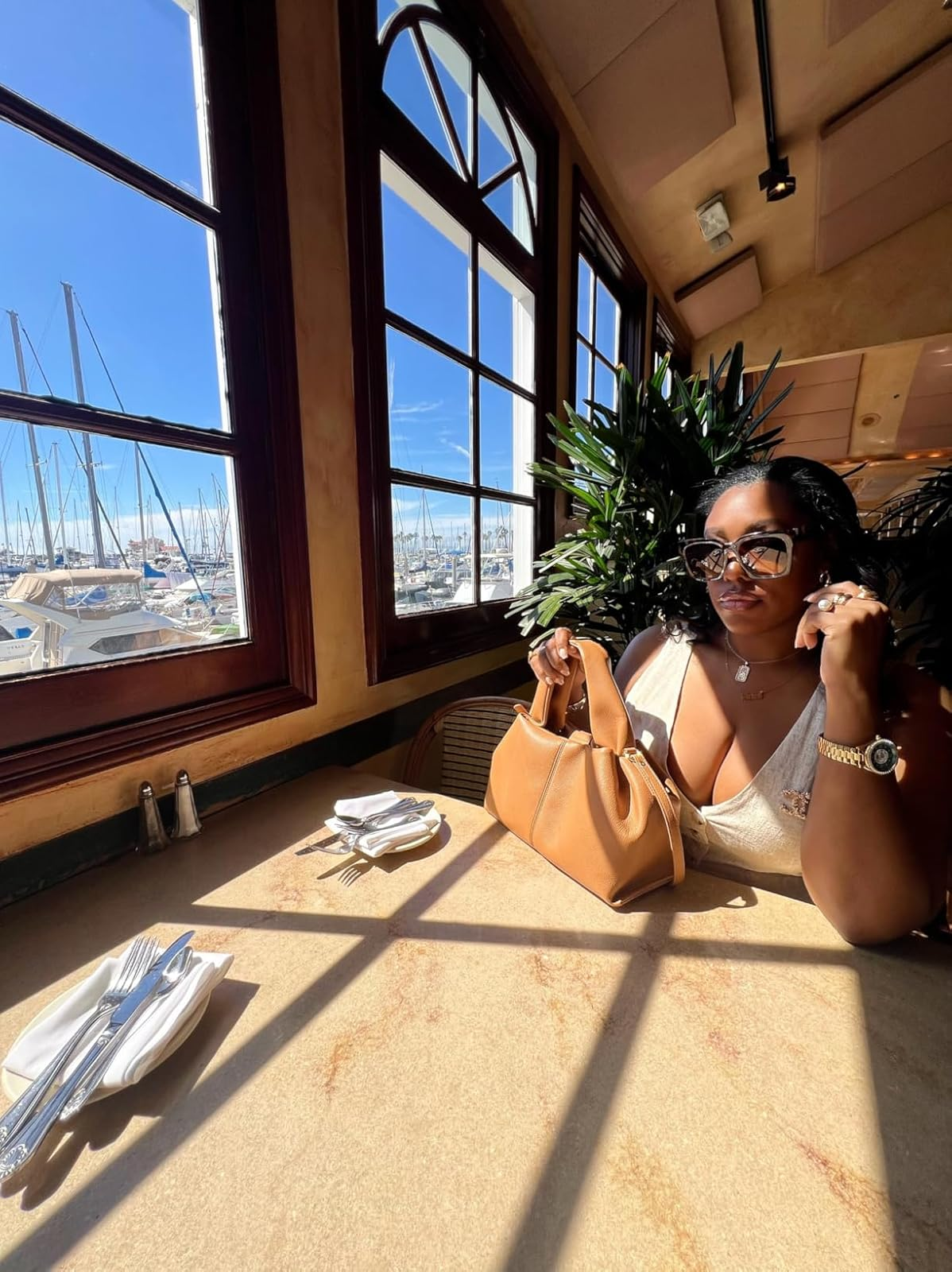 Person in sunglasses and a chic white top, sitting at a sunlit table near marina view, holding a tan bag, ready for a casual outdoor dining experience