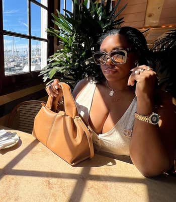 Person in sunglasses and a chic white top, sitting at a sunlit table near marina view, holding a tan bag, ready for a casual outdoor dining experience
