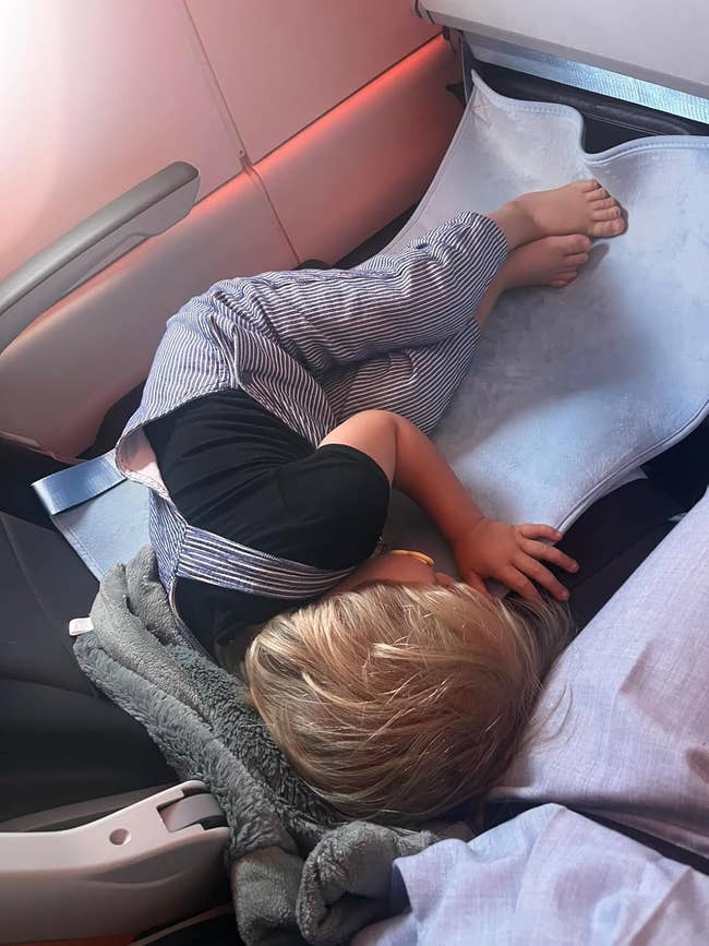 A child in striped overalls and a dark shirt sleeps on a makeshift bed on an airplane seat 