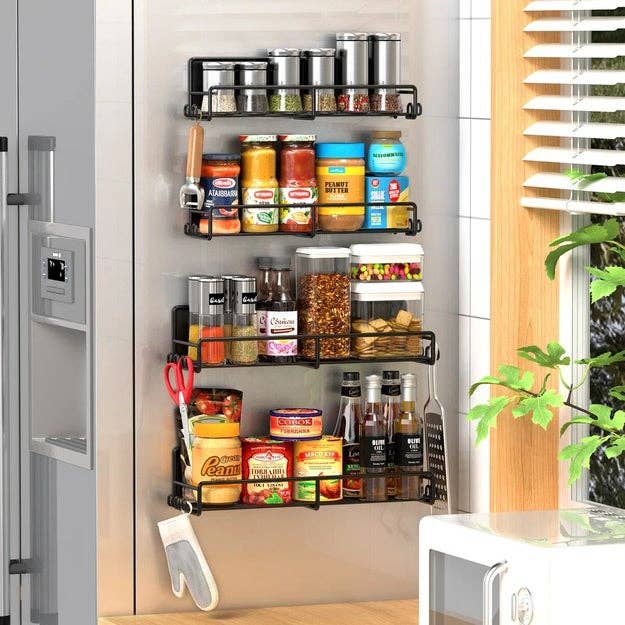 Wall-mounted spice rack in kitchen, holding various jars and containers of spices and condiments, next to a window with greenery outside
