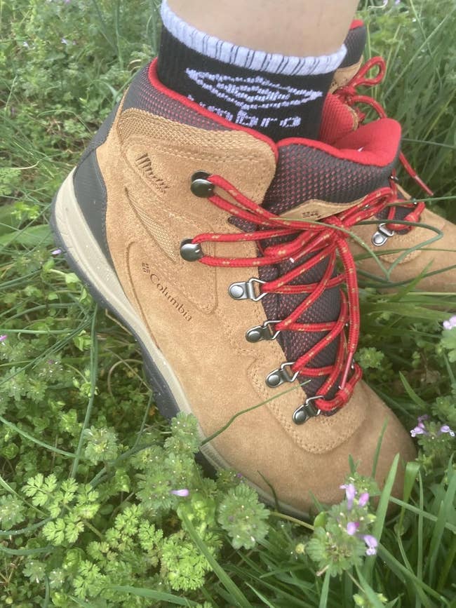 A close-up of a tan hiking footwear with reddish laces and achromatic trim connected grass, worn with a achromatic sock