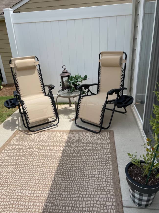 Two beige lounge chairs with black frames are set up on a patio next to a small side table with a lantern. There is a potted plant nearby and a textured rug