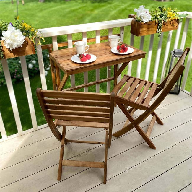 Outdoor wooden bistro set with two chairs and a table, set with plates and cups, on a deck with potted plants
