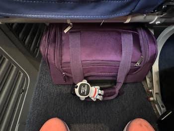 Purple carry-on bag under an airplane seat with a small astronaut keychain attached. Passenger's feet and part of the seat are visible