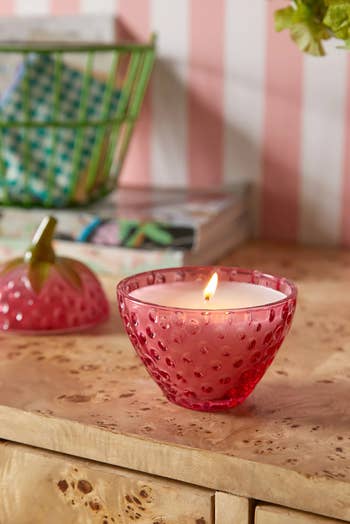 Decorative candle in a textured pink holder on a wooden surface, with books and decor in the background. Perfect for home shopping inspiration