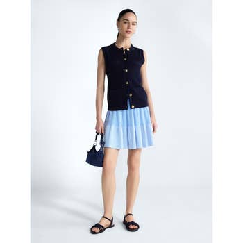 Person wearing a sleeveless button-up top, pleated skirt, sandals, and holding a small handbag, posing as in a fashion catalog