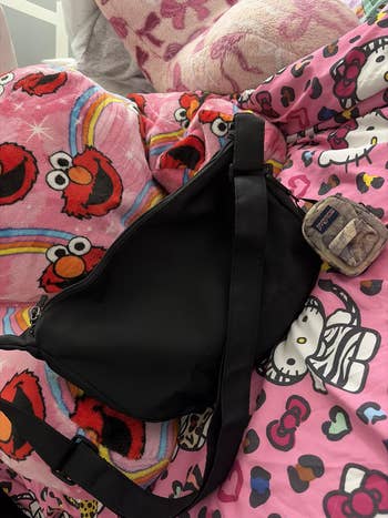 A black crossbody bag sits on a colorful bedspread featuring cartoon characters like Elmo and Hello Kitty. A small camo pouch is next to it