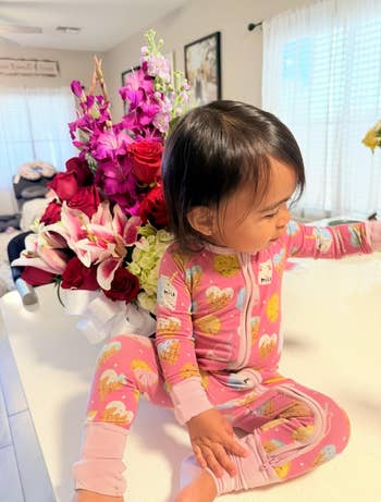 A toddler successful patterned pajamas sits connected a countertop adjacent to a ample bouquet of flowers indoors